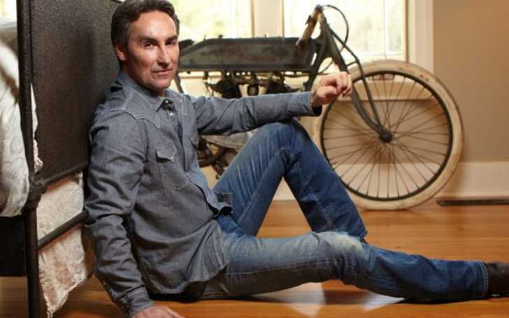 Picture of Mike Wolfe posing for a photo inside one of his luxurious house with old school cycle in backgraound.